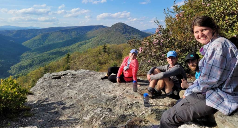 A group of people sit on a rock overlook. Below them is a vast green mountainous area. 